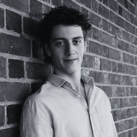 Black and white portrait of a young man with dark hair wearing a light button-up shirt, standing against a brick wall